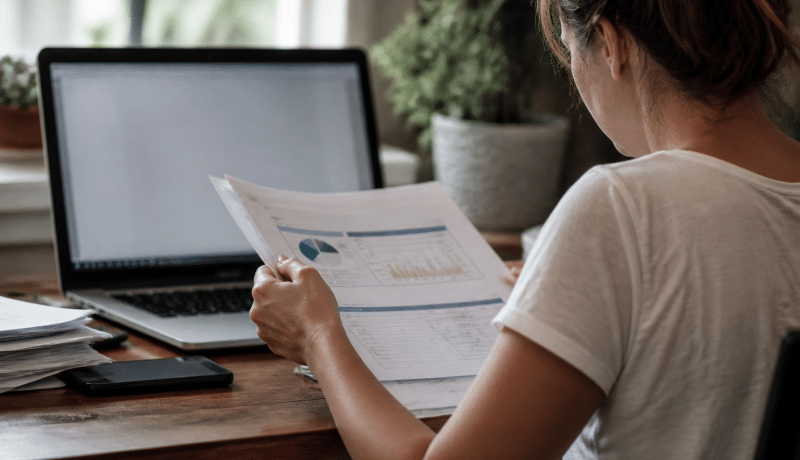 Personne assise devant ordinateur consultant documents financiers papier.