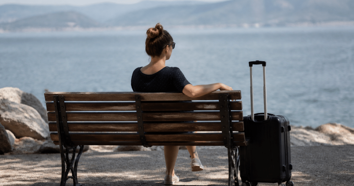 Personne assise sur un banc, valise à côté, regardant vers l'horizon.