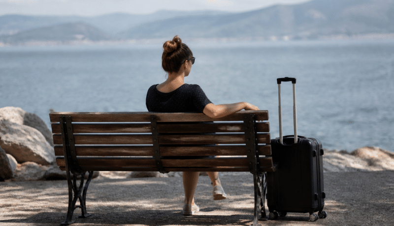 Personne assise sur un banc, valise à côté, regardant vers l'horizon.