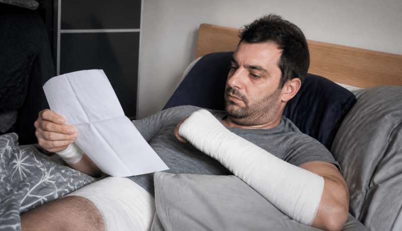 Travailleur immobilise au lit avec pansements et documents administratifs.